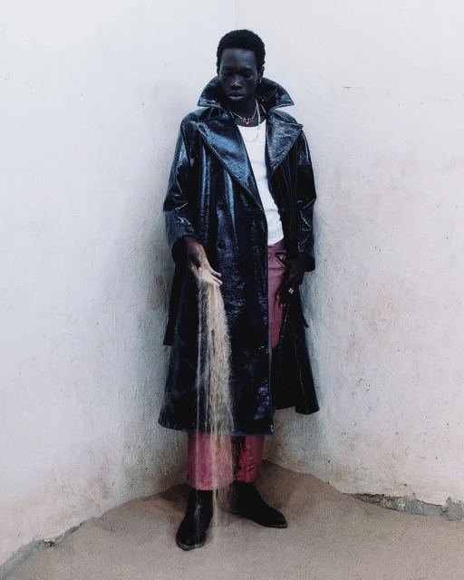 Dark-skinned adult male in a black patent-leather trench coat holds a stream of sand against a white wall in an indoor corner setting.