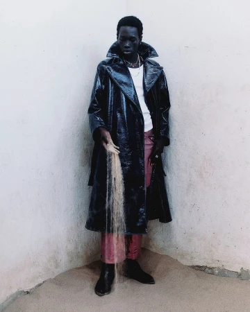 Dark-skinned adult male in a black patent-leather trench coat holds a stream of sand against a white wall in an indoor corner setting.