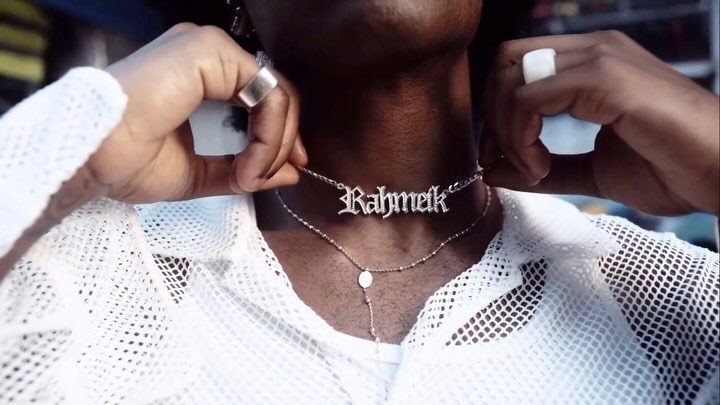 Close-up of a person wearing two silver necklaces, one with the name 'Rahmelk,' and a white fishnet shirt with a white undershirt.