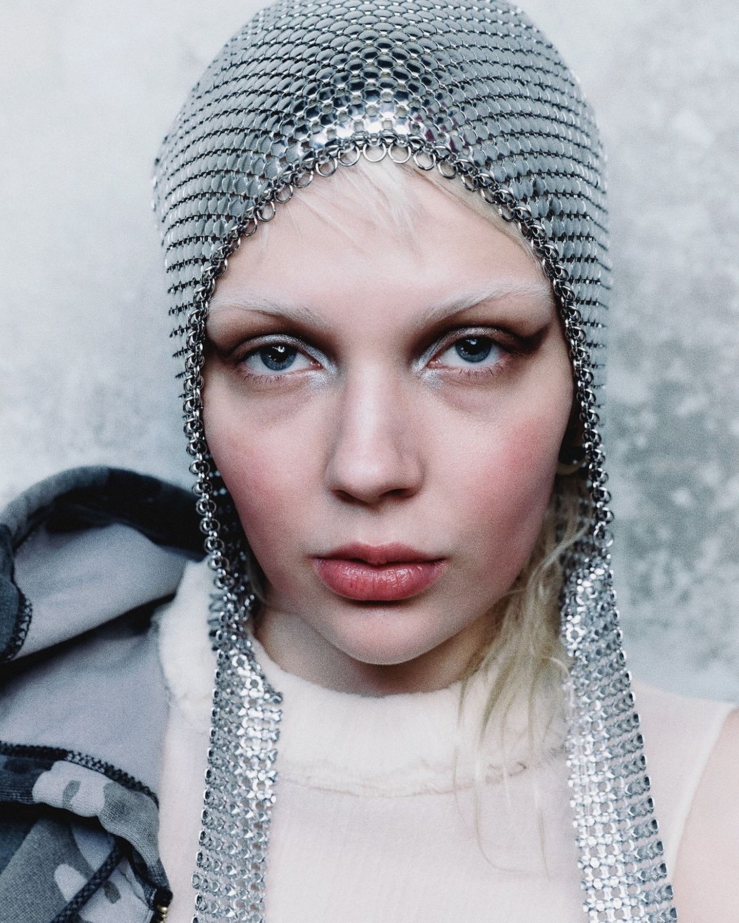Close-up portrait of a fair-skinned person with a metallic chainmail head covering, light blonde hair, and pale pink makeup.