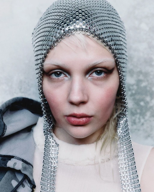 Close-up portrait of a fair-skinned person with a metallic chainmail head covering, light blonde hair, and pale pink makeup.