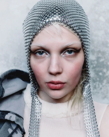 Close-up portrait of a fair-skinned person with a metallic chainmail head covering, light blonde hair, and pale pink makeup.