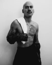 Portrait of a tattooed bald man with a mustache wearing dark gloves and dark trousers against a white background