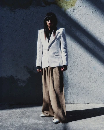 Model with long dark hair wearing a white structured blazer and tan trousers standing outdoors.