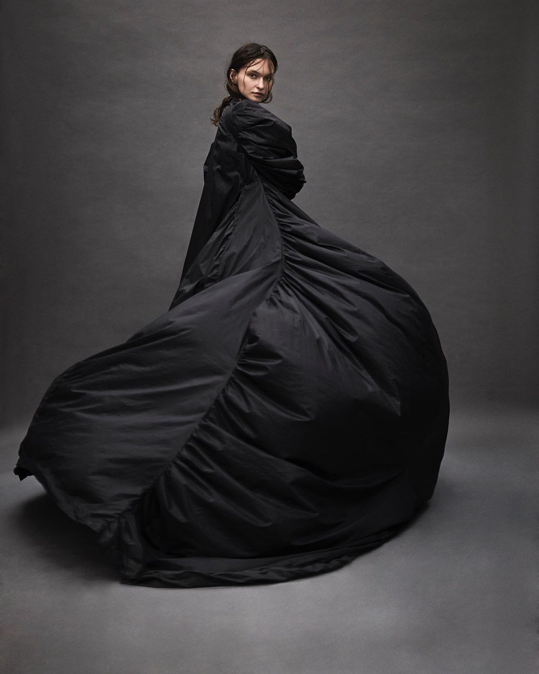 A slender woman in a flowing black gown gazes over her shoulder in a studio shot, exhibiting a blend of fashion and fine art photography.