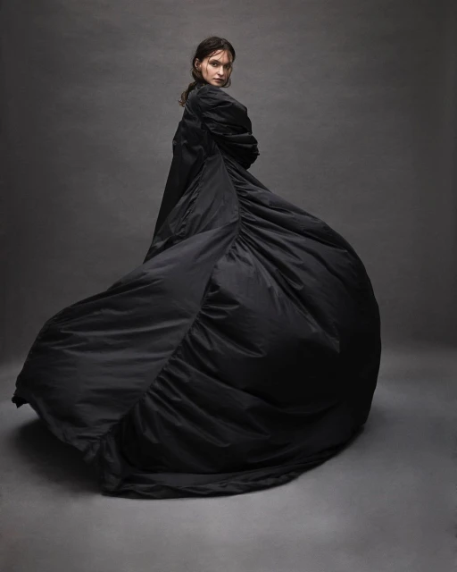 A slender woman in a flowing black gown gazes over her shoulder in a studio shot, exhibiting a blend of fashion and fine art photography.