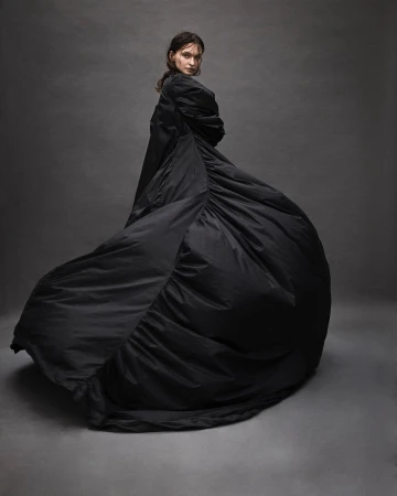 A slender woman in a flowing black gown gazes over her shoulder in a studio shot, exhibiting a blend of fashion and fine art photography.