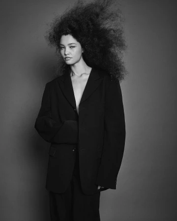 Portrait of a woman with voluminous dark hair wearing an oversized black blazer against a grey backdrop.