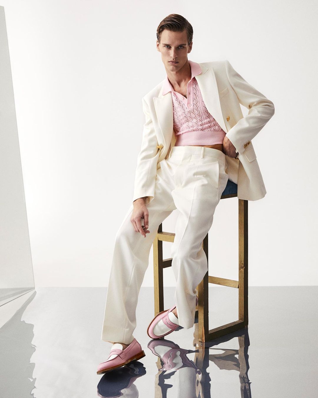 A young adult man is posed in a light-pink top and off-white suit while sitting on a stool in a bright, reflective studio environment.