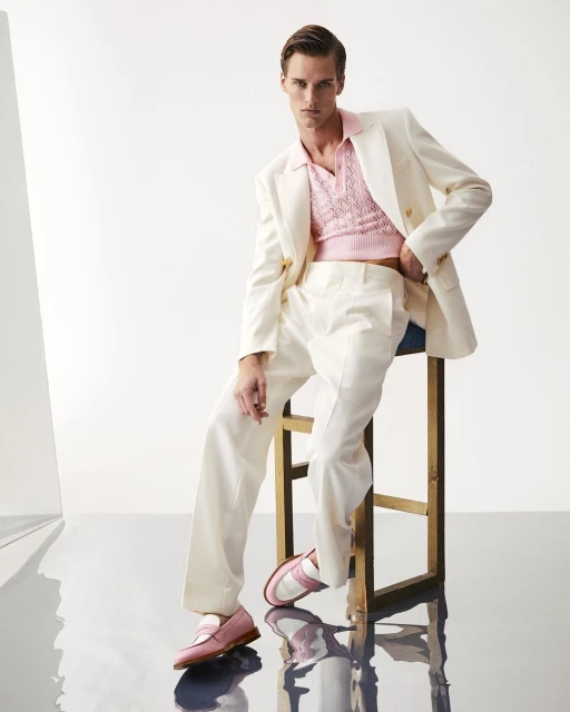 A young adult man is posed in a light-pink top and off-white suit while sitting on a stool in a bright, reflective studio environment.