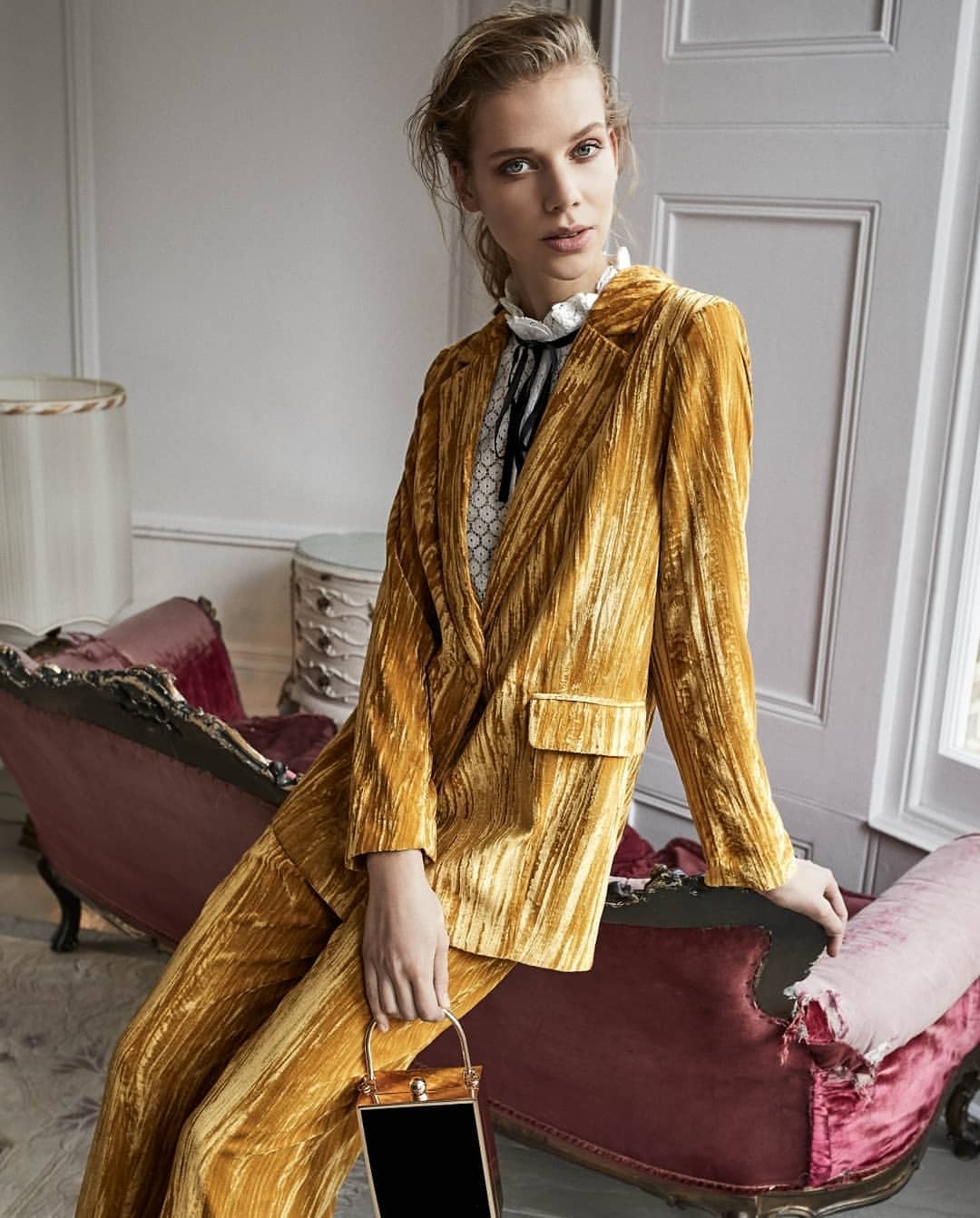 Woman in golden yellow crushed velvet suit with black purse poses indoors on red velvet chaise lounge looking at the camera.