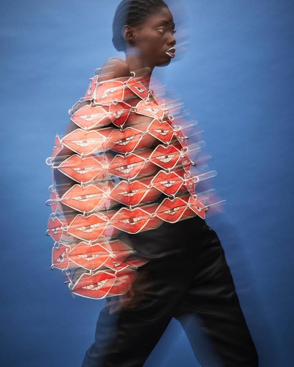 Profile of a Black female model with white lipstick wearing a red lips top and black pants against a blue background in motion blur.