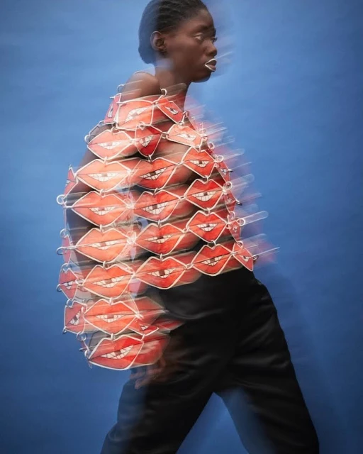 Profile of a Black female model with white lipstick wearing a red lips top and black pants against a blue background in motion blur.