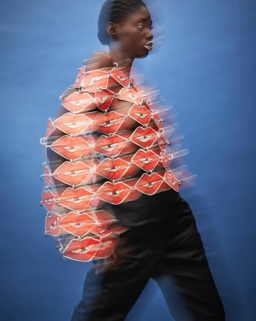 Profile of a Black female model with white lipstick wearing a red lips top and black pants against a blue background in motion blur.