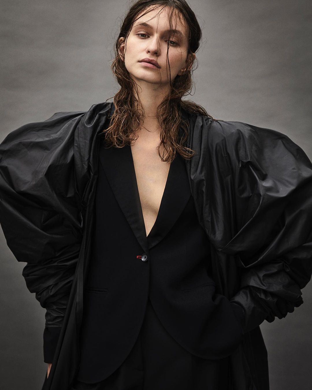 Portrait of a woman with wet hair wearing a black blazer with dramatic voluminous sleeves against a textured grey backdrop.