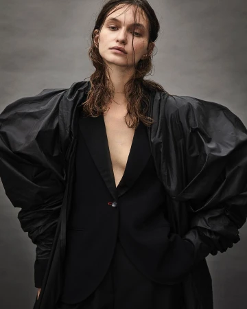 Portrait of a woman with wet hair wearing a black blazer with dramatic voluminous sleeves against a textured grey backdrop.