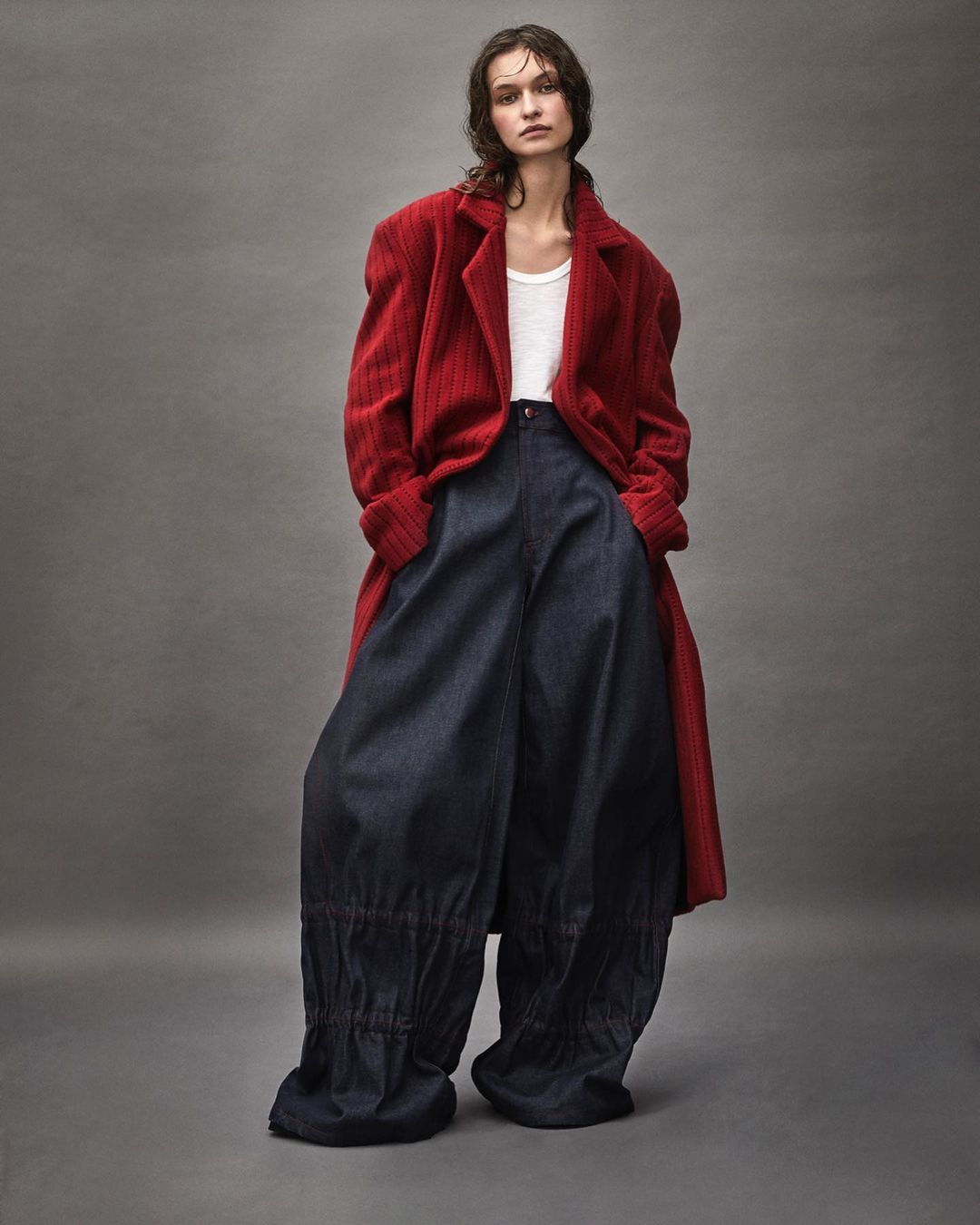A woman poses in a red overcoat, white tank top, and dark blue baggy pants against a neutral grey backdrop in a fashion editorial studio shot.