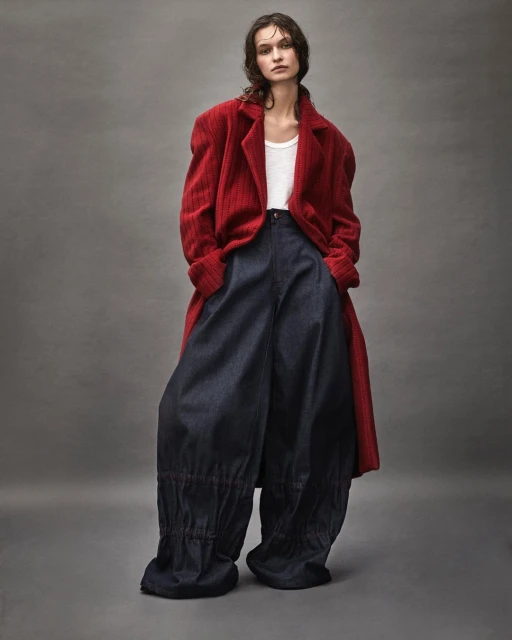 A woman poses in a red overcoat, white tank top, and dark blue baggy pants against a neutral grey backdrop in a fashion editorial studio shot.