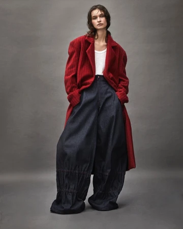 A woman poses in a red overcoat, white tank top, and dark blue baggy pants against a neutral grey backdrop in a fashion editorial studio shot.