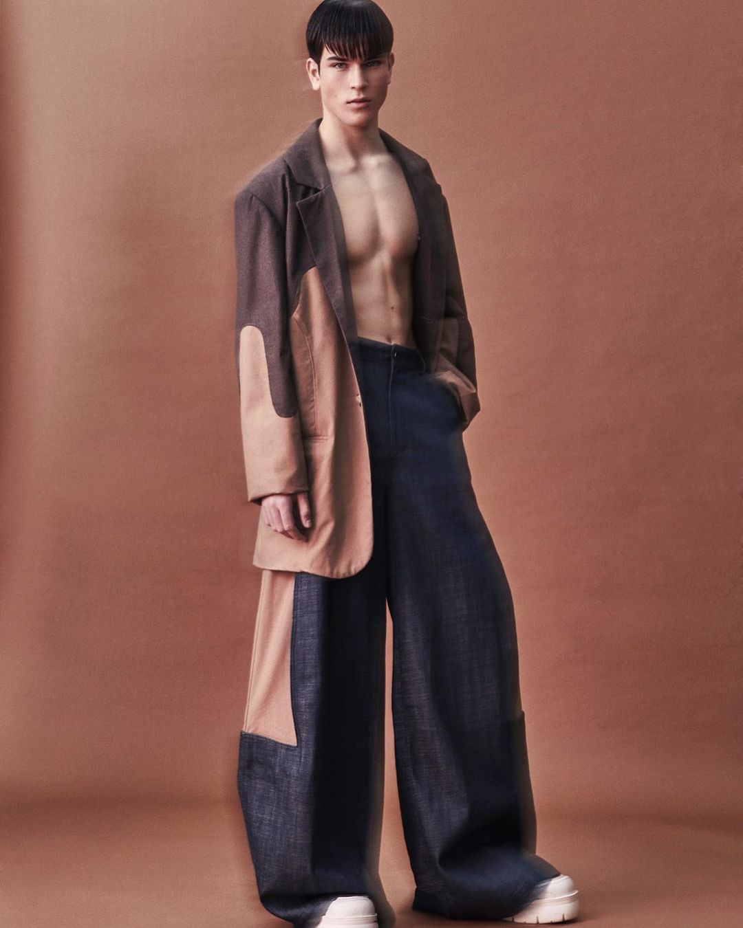 Full-length shot of an adult male fashion model in a brown and beige oversized coat with blue flared pants against a brown background.