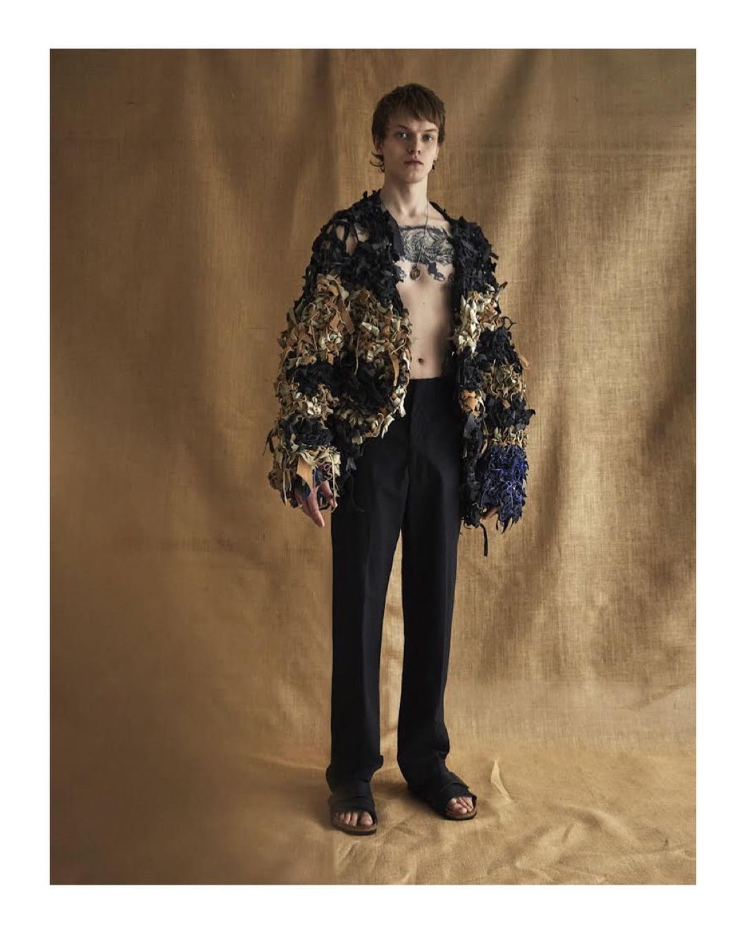 Full-length portrait of a young adult male wearing a textured jacket, dark trousers, and sandals on a brown paper background.