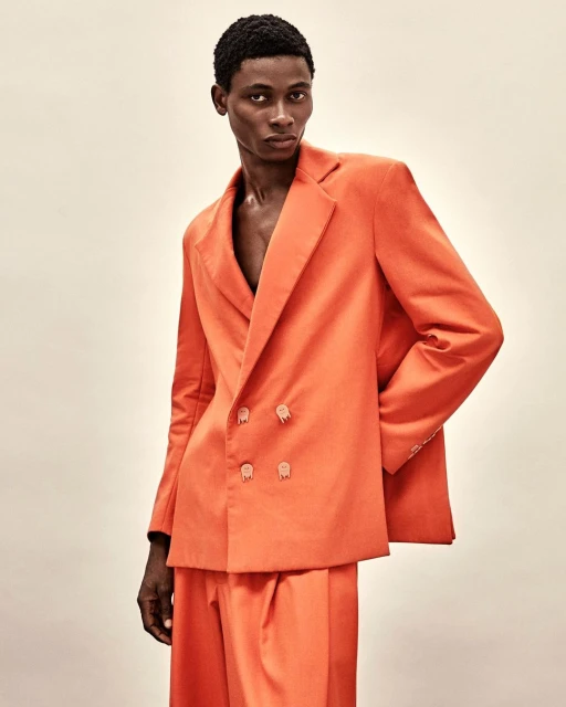 Portrait of a young Black male model in an orange double-breasted suit, posing indoors against a neutral backdrop in a fashion editorial.