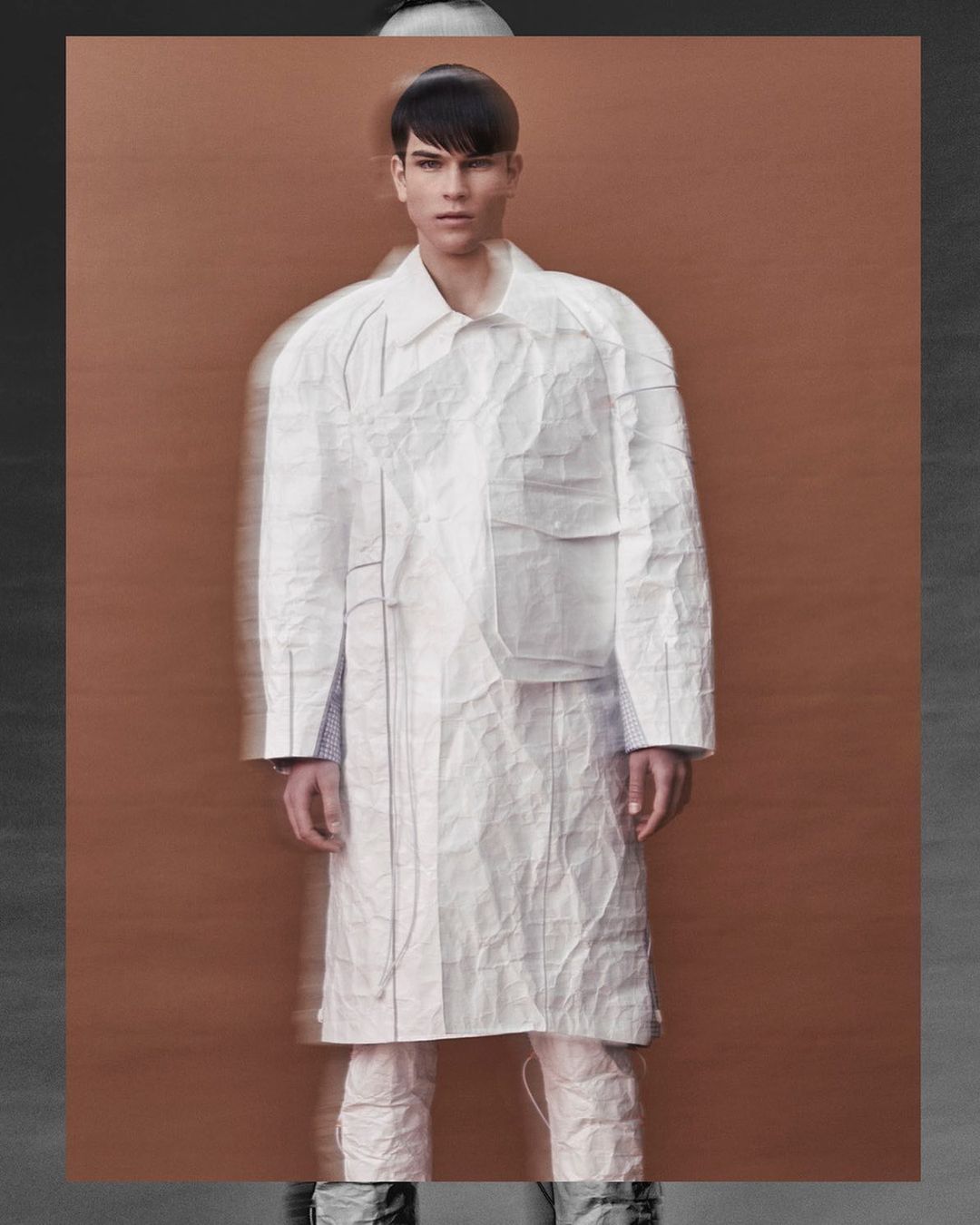 Full shot of male fashion model in a white paper-like textured coat and matching pants against a tan and gray background.