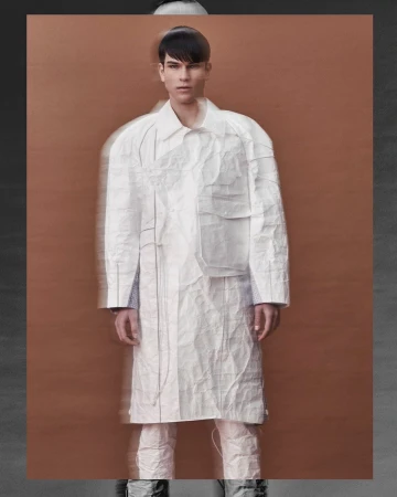 Full shot of male fashion model in a white paper-like textured coat and matching pants against a tan and gray background.
