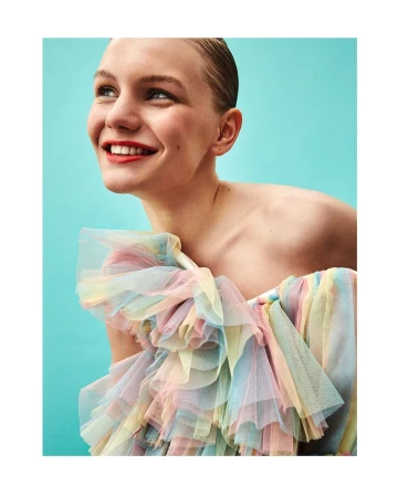 Smiling woman with a rainbow tulle gown poses against a bright teal background in a fashion portrait with a shallow depth of field.