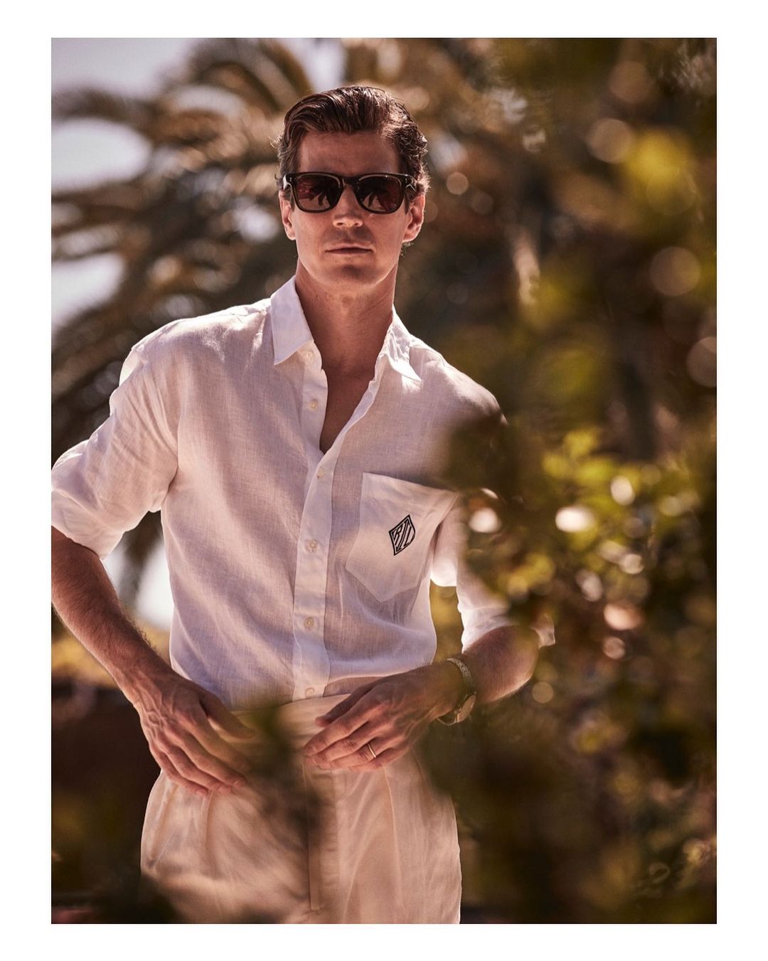 A man in a white shirt and tan pants wearing sunglasses stands outdoors with a palm tree and foliage in the background.