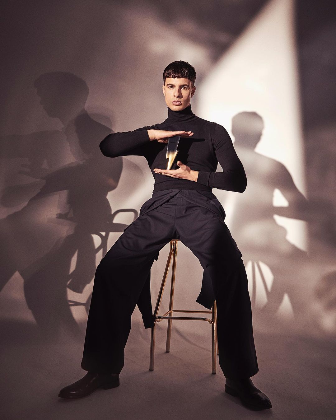 An adult male sits on a gold stool, wearing a black turtleneck and wide-legged pants, holding a lightning bolt object in a studio.
