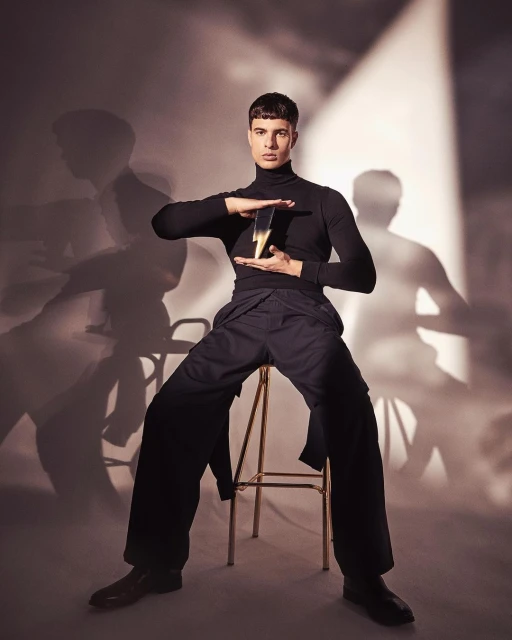 An adult male sits on a gold stool, wearing a black turtleneck and wide-legged pants, holding a lightning bolt object in a studio.