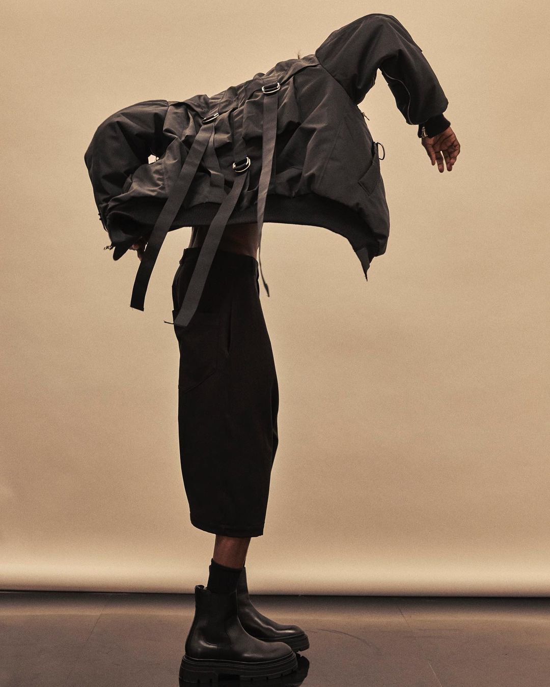 Model wears a dark grey bomber jacket with straps and buckles paired with black culottes and Chelsea boots, posing in a studio setting.
