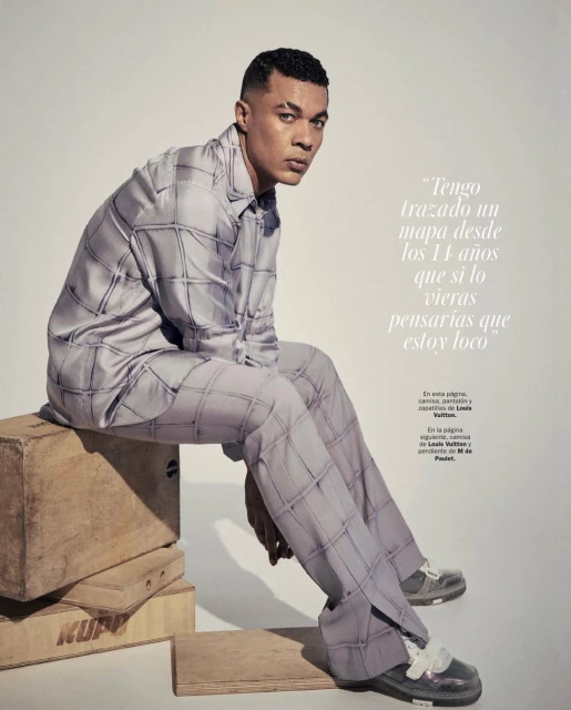 Man in patterned pajamas sitting on wooden boxes in a studio. He looks at the camera with a neutral expression against a plain background.