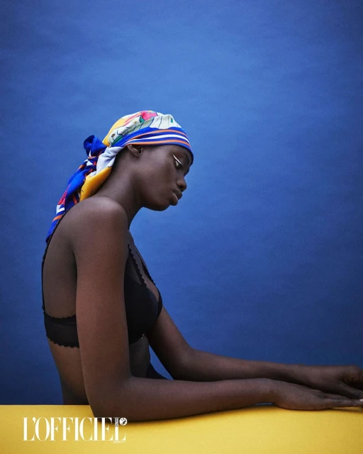 Portrait of a Black woman wearing a patterned headscarf and black lingerie against a solid blue background in a studio setting.