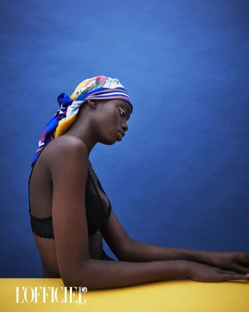 Portrait of a Black woman wearing a patterned headscarf and black lingerie against a solid blue background in a studio setting.