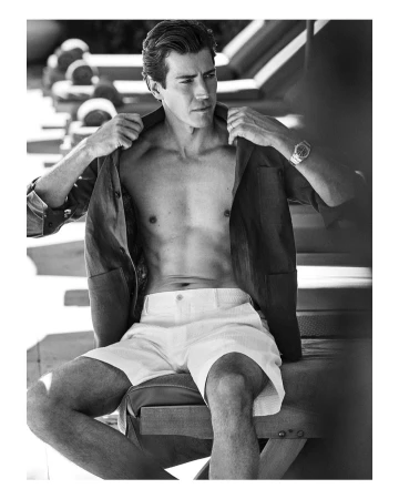 Monochrome portrait of a man sitting outdoors adjusting an open shirt while wearing light shorts and a wrist watch.