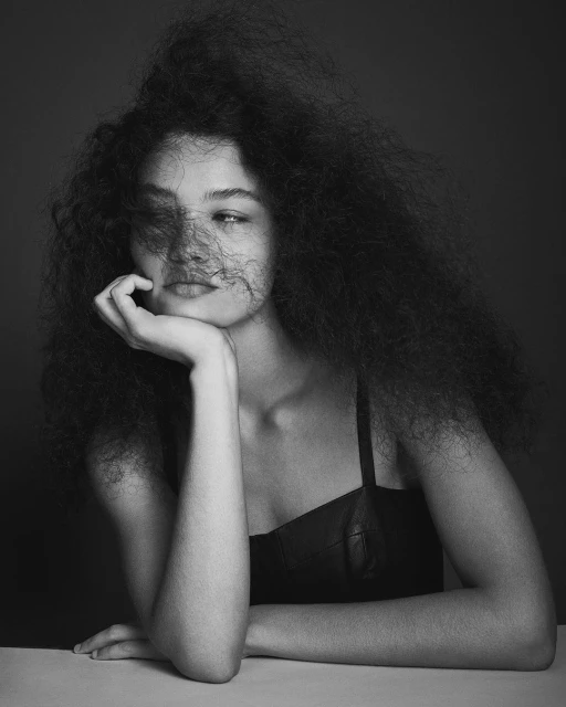 Black-and-white portrait of a woman with large, curly hair resting her chin on her hand and leaning on a table in a studio setting.