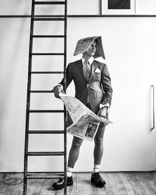Black and white fashion shot of a man in a suit and newspaper hat standing next to a ladder, holding a newspaper.