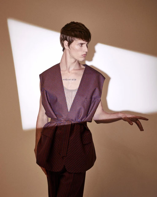 A fair-skinned young adult male model poses in a purple pinstripe outfit against a neutral background in a fashion shot with directional lighting.