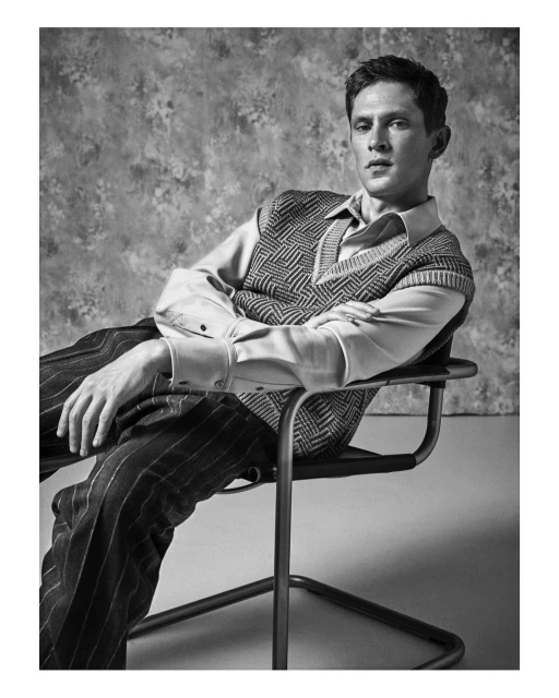 Black and white portrait of a young adult man in a patterned vest, sitting on a chair in a studio setting, looking directly at the camera.
