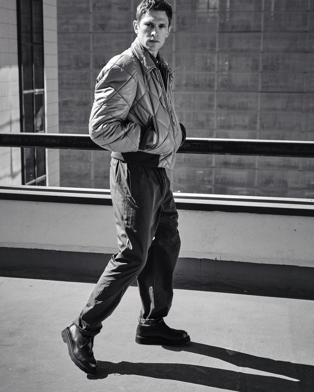 Monochrome full shot of an adult male in a quilted jacket and dark pants walking on a concrete surface, with an urban backdrop.