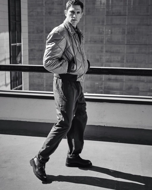 Monochrome full shot of an adult male in a quilted jacket and dark pants walking on a concrete surface, with an urban backdrop.