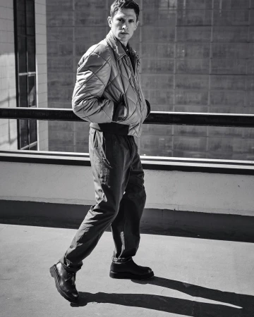Monochrome full shot of an adult male in a quilted jacket and dark pants walking on a concrete surface, with an urban backdrop.