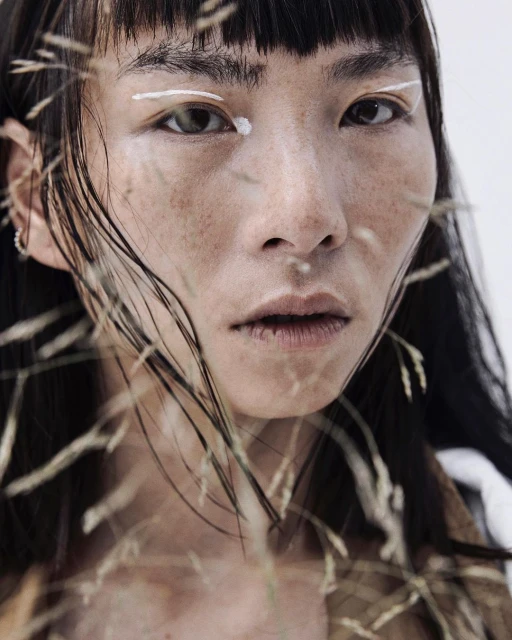 Close-up portrait of an Asian female model with fair skin and dark hair, showcasing white eyeliner and surrounded by wheat stalks.