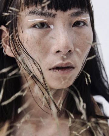 Close-up portrait of an Asian female model with fair skin and dark hair, showcasing white eyeliner and surrounded by wheat stalks.