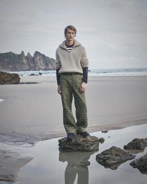 A fair-skinned male model wearing a knit sweater and geometric quilted pants stands on a rock on a beach with cliffs in the background.