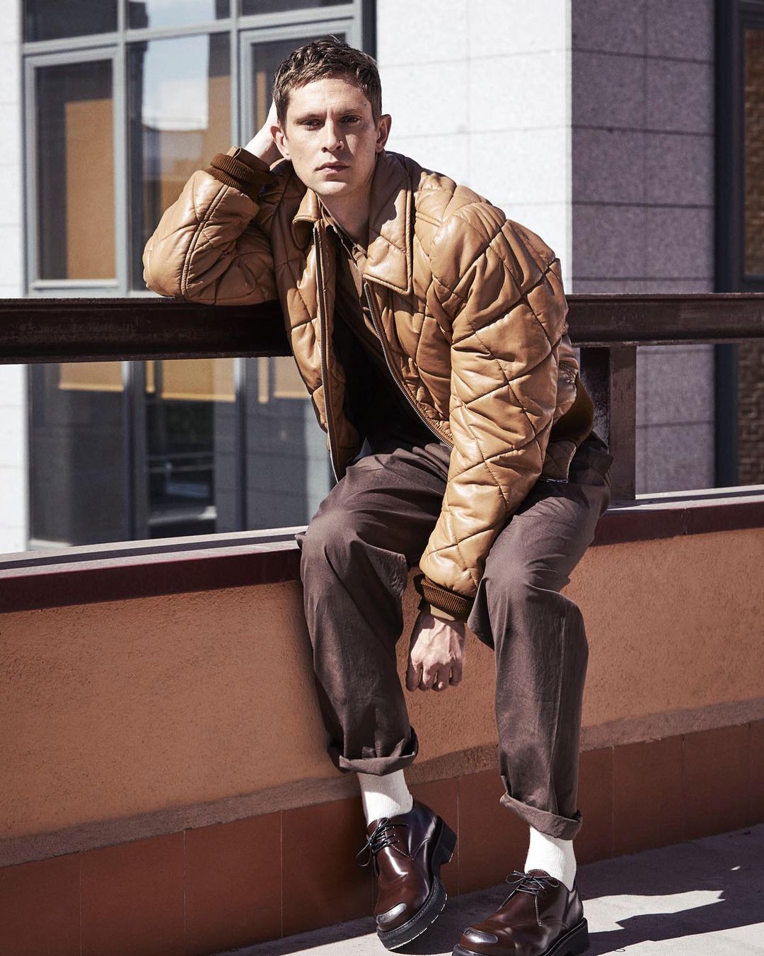 A young adult man poses seated outdoors in a brown quilted jacket, brown trousers, and polished dark brown leather shoes with thick soles.