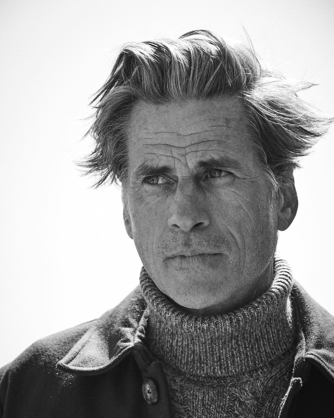 Black and white portrait of a mature adult male in a textured sweater and jacket looking away from the camera with a serious expression.