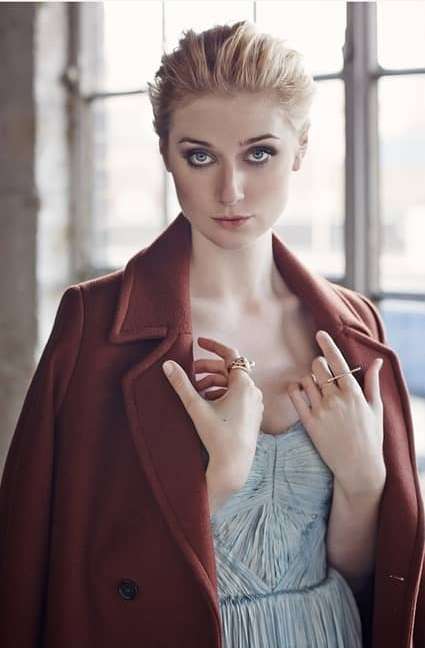 Blonde woman in a rust-colored coat poses indoors, wearing rings and a light blue dress, near a window, with medium-shot framing.
